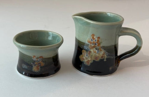 A lovely little cream and sugar set handmade in ceramic.  Colors include celadon, orange, black and brown.  It has small floral details in shades of orange added to each.  The top half is celadon and the other colors are layered on the bottom half.  The cream pitcher has a little handle but the sugar bowl does not.  A very beautiful, functional addition to your table.