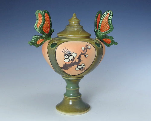 A beautiful lidded ceramic pedestal jar with two hand fabricated mounted,large butterflies - one on each side.  The butterflies are green and orange and the jar is mostly green with peach, brown and white, as well.  There are white flowers on a brown branch painted on the front.  The lid is pointed at the top and the main body of the piece is rounded on a slim pedestal base.