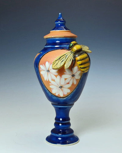 A wonderful ceramic lidded blue pedestal jar with terracotta color sections that have white flowers painted on them. It has a large, hand fabricated, yellow and black bee mounted on the side right below the lid.  The lid is tapered to a point on top of a rounded body jar on a slim pedestal base.  